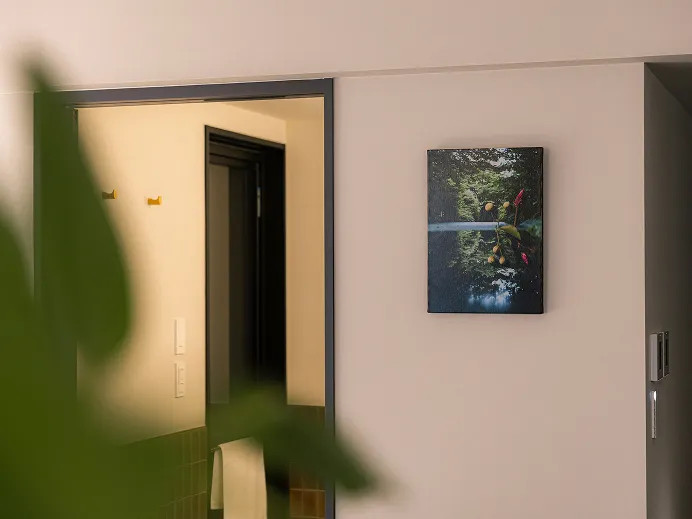 開いた木製の扉と額装された植物の写真が見える玄関まわり。手前に大きな観葉植物の葉がのぞく。